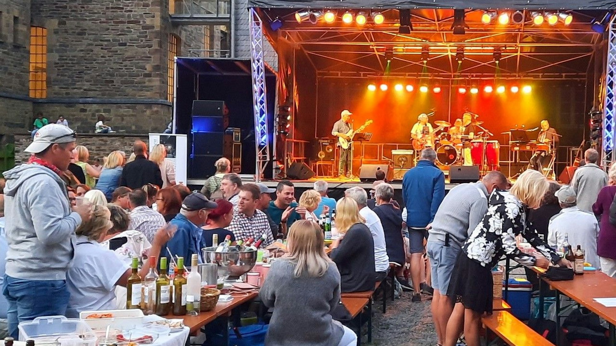 Eine Open-Air-Bühne. Davor sitzen Menschen auf Bierzeltbänken an Tischen.