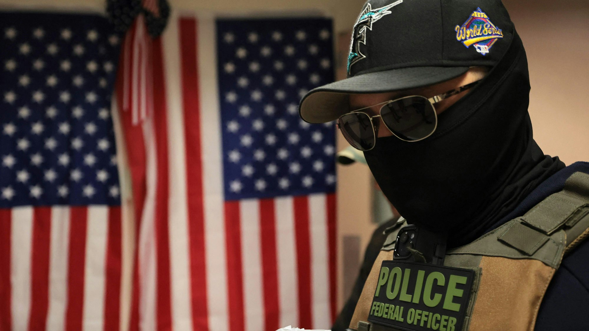 Ein vermummter US-Bundesbeamter mit Kappe, Sonnenbrille und Weste steht vor einer amerikanischen Flagge.