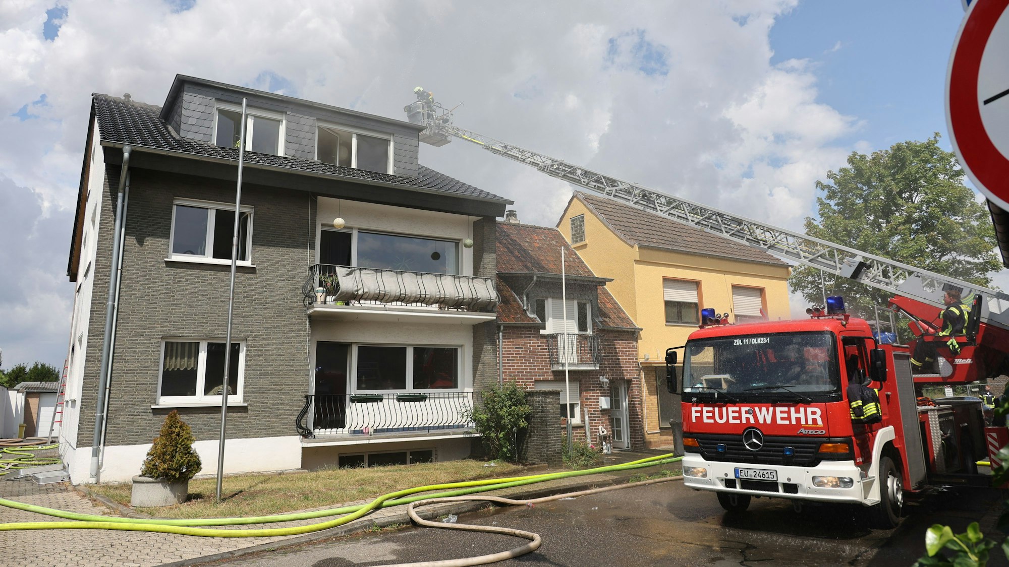 Die Drehleiter ist vor dem Haus, in dem ein Feuer ausgebrochen ist, in Stellung gegangen. Oben im Korb öffnet ein Feuerwehrmann mit einem Einreißhaken die Abdeckung des Dachs.
