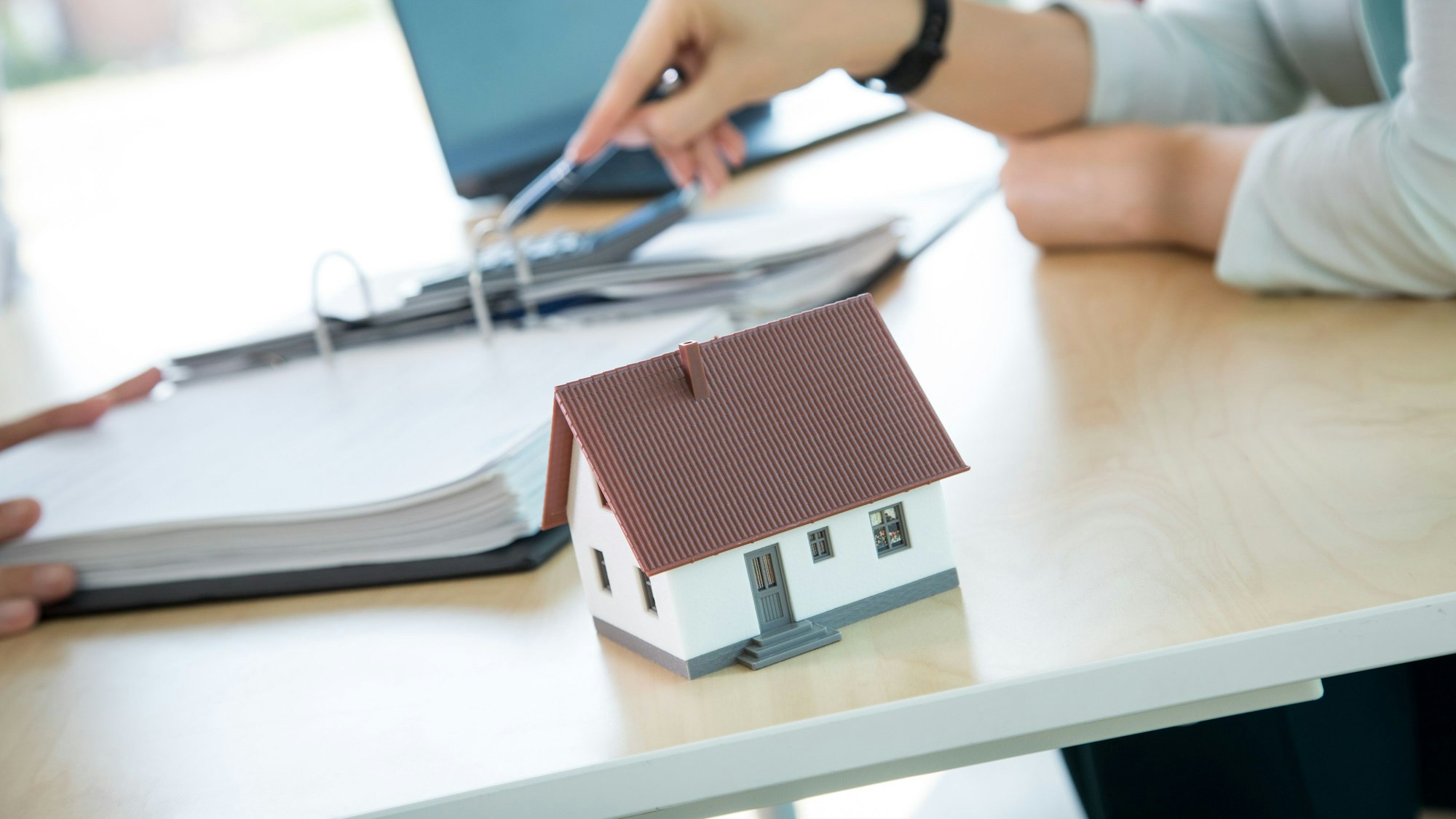 Ein Modellhaus steht auf einem Schreibtisch, im Hintergrund deutet eine Hand auf Unterlagen.