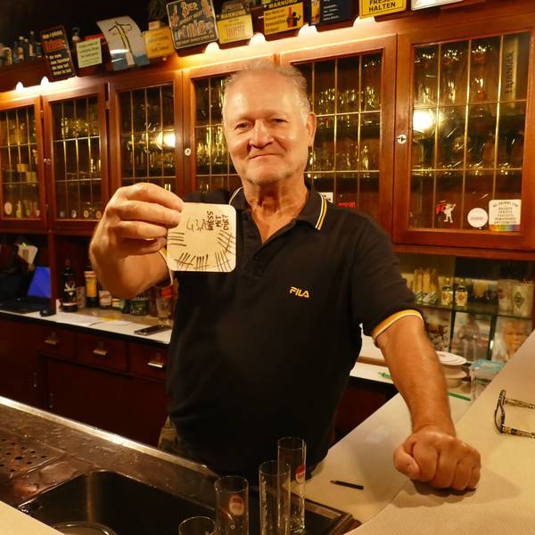 Wolfgang Ort hält einen voll beschriebenen Bierdeckel in der Hand. Er steht hinter der Theke seiner Kneipe.