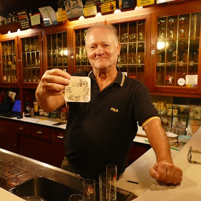 Wolfgang Ort hält einen voll beschriebenen Bierdeckel in der Hand. Er steht hinter der Theke seiner Kneipe.