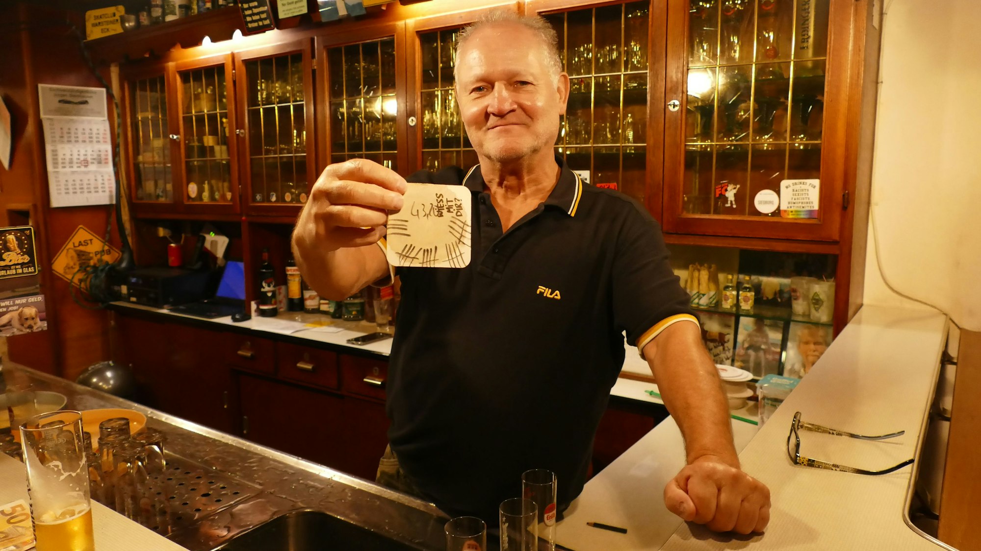 Wolfgang Ort hält einen voll beschriebenen Bierdeckel in der Hand. Er steht hinter der Theke seiner Kneipe.