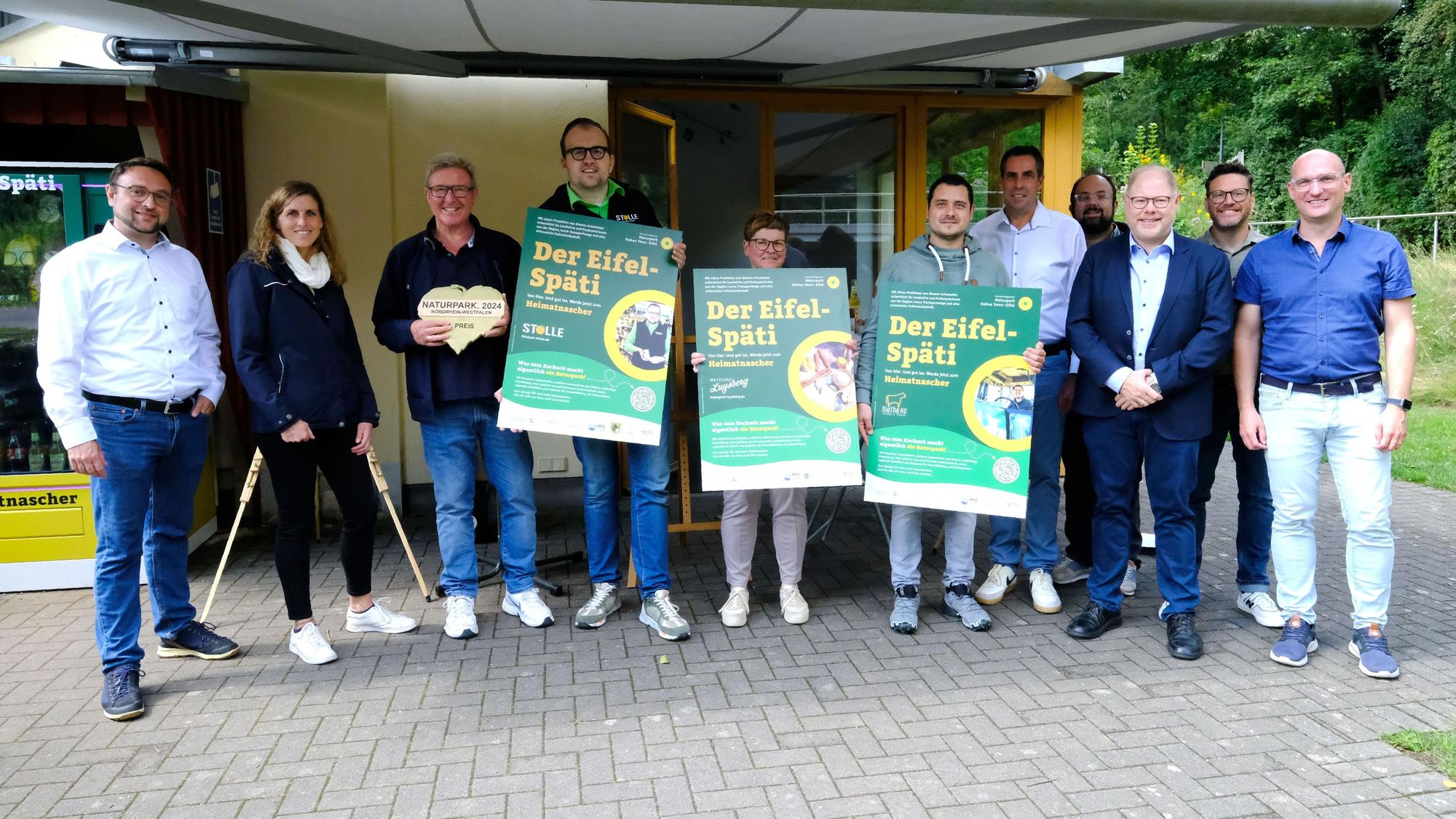 Gruppenbild der Projektbeteiligten und Automatenbetreiber. Drei Personen halten Plakate mit der Aufschrift „Der Eifel-Späti“.