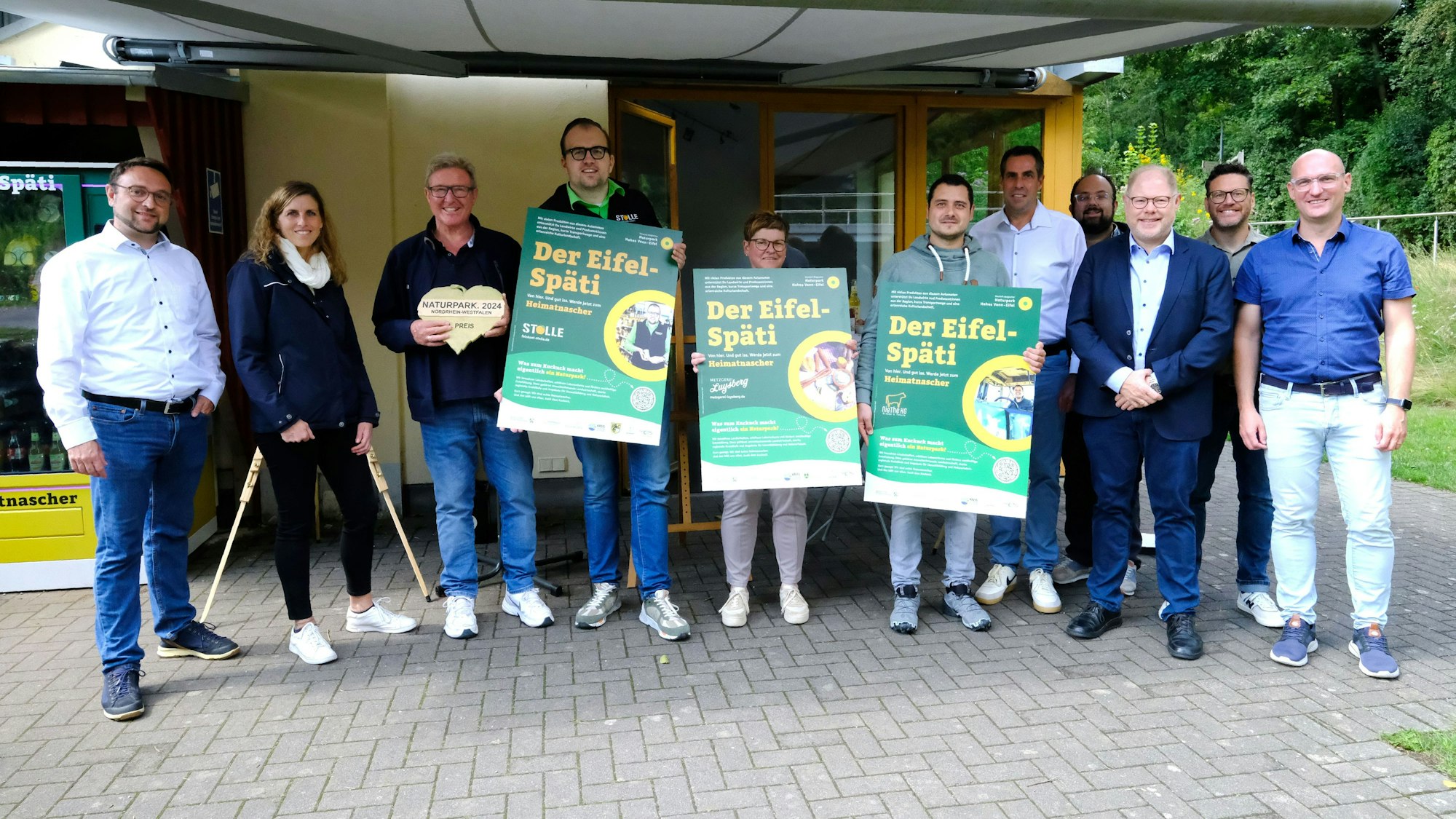 Gruppenbild der Projektbeteiligten und Automatenbetreiber. Drei Personen halten Plakate mit der Aufschrift „Der Eifel-Späti“.