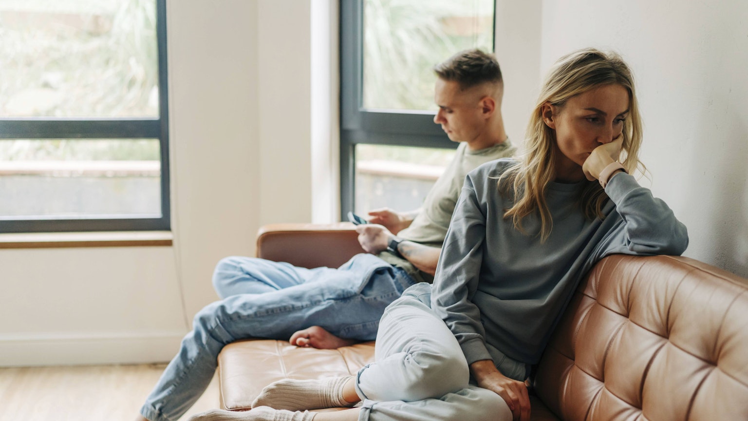 Eine junge Frau ist traurig und beleidigt von ihrem Partner. Der sitzt neben ihr auf der Couch und schaut auf sein Handy.