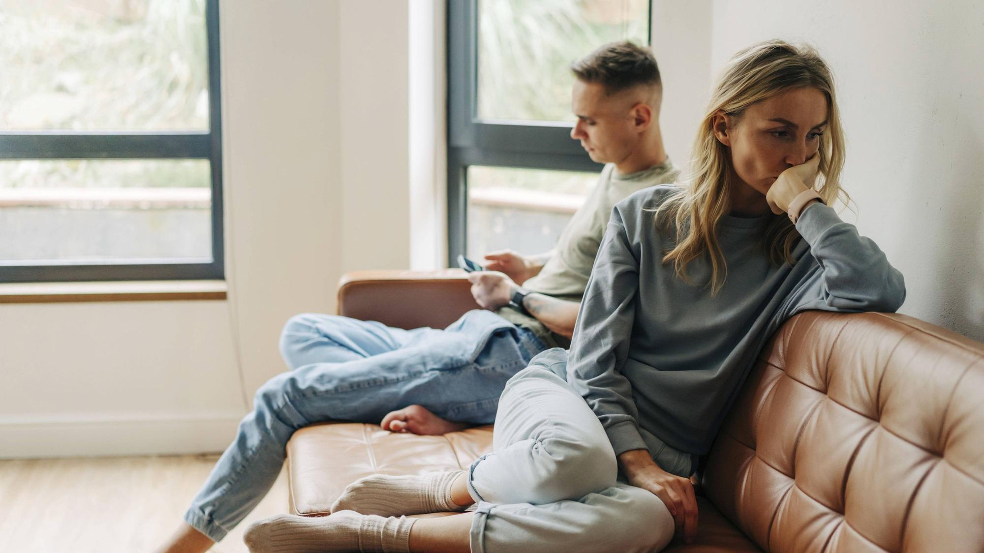 Eine junge Frau ist traurig und beleidigt von ihrem Partner. Der sitzt neben ihr auf der Couch und schaut auf sein Handy.