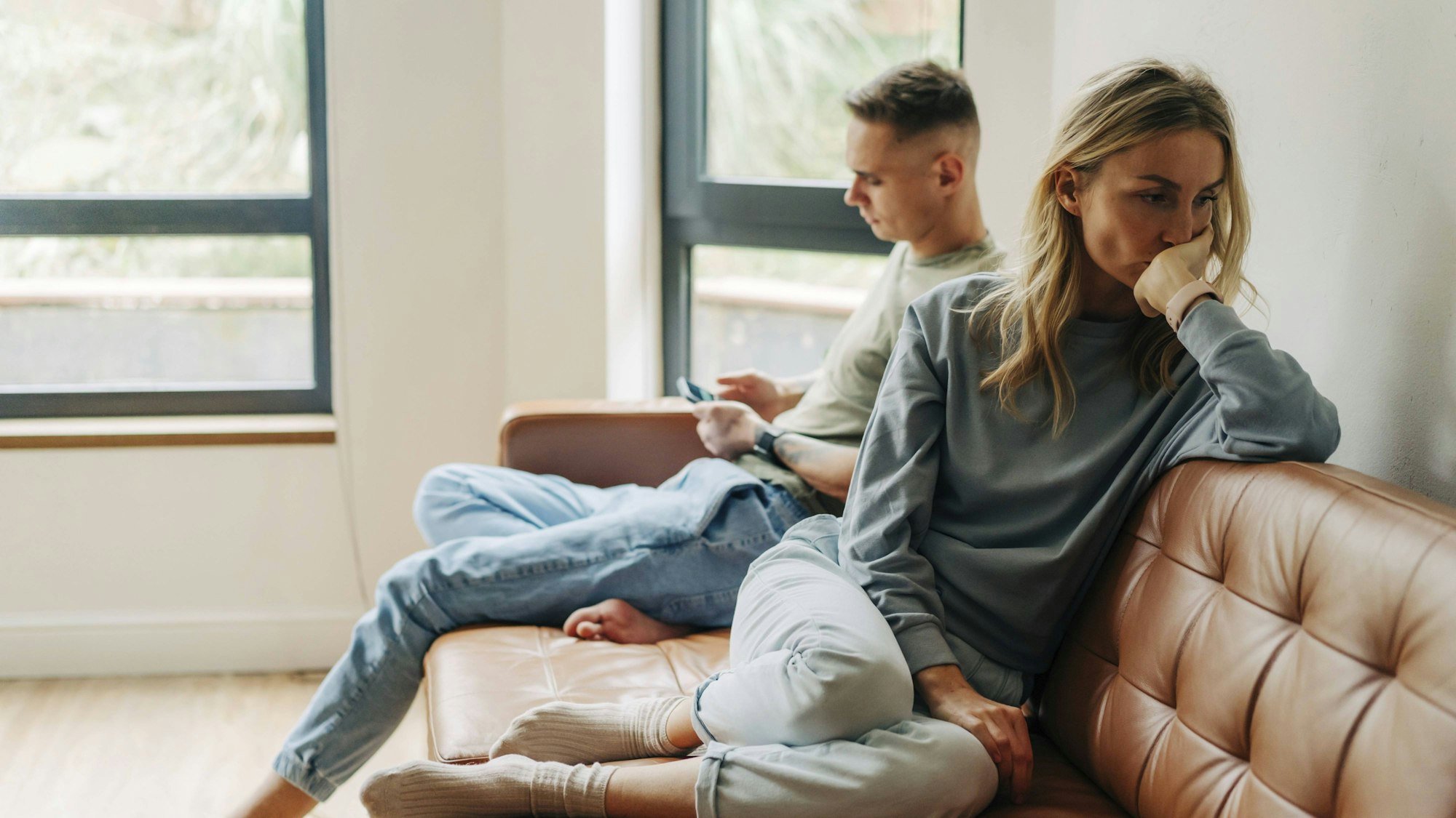 Eine junge Frau ist traurig und beleidigt von ihrem Partner. Der sitzt neben ihr auf der Couch und schaut auf sein Handy.