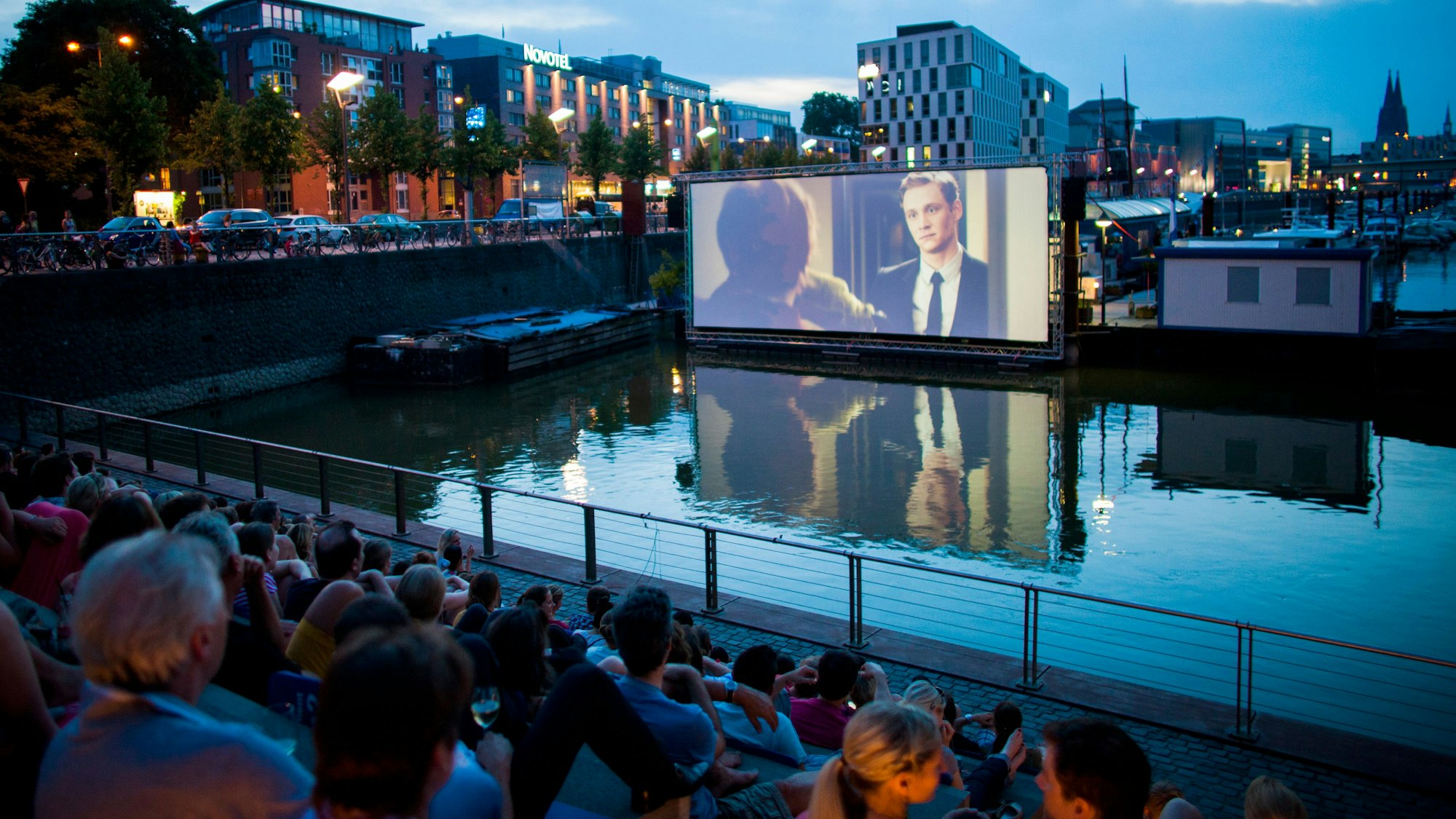 Menschen schauen sich im Open Air Kino am Rheinauhafen in Köln vor der Domkulisse einen Film an. (Archivbild)