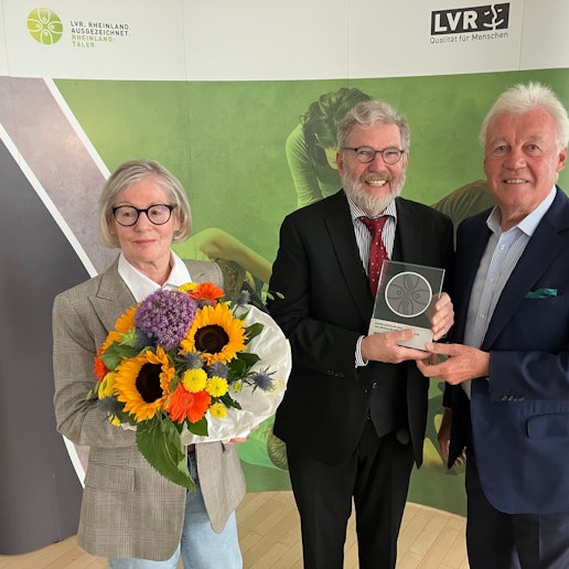 Eine Dame mit Blumenstrauß, zwei Herren mit einer gerahmten Medaille.