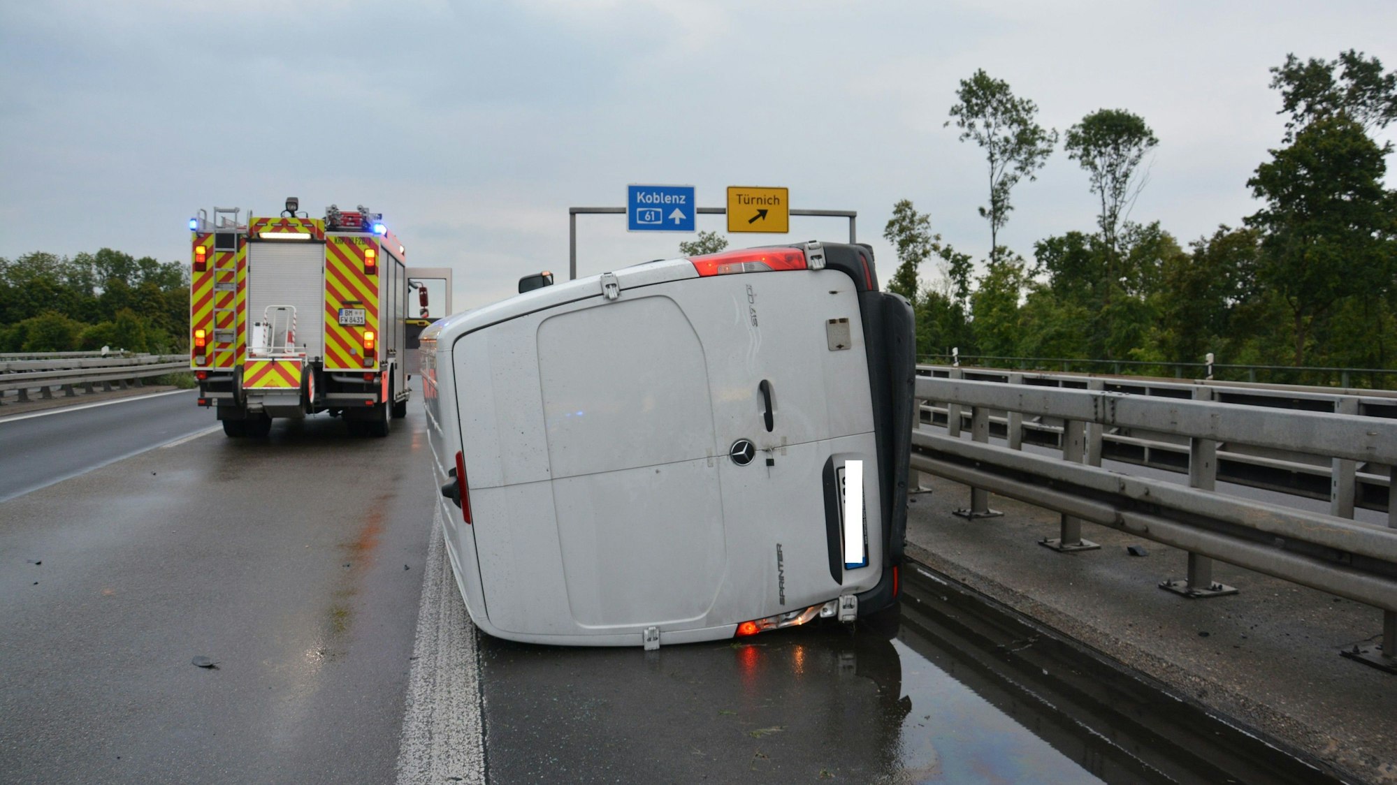 Ein Sprinter liegt auf der Seite. Im Hintergrund die Schilder der A61 und der Ausfahrt Türnich. Ein Feuerwehrfahrzeug ist zu sehen.