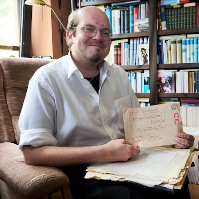 Der Autor Andreas Züll sitzt vor einem Regal voller Bücher und hält einen alten Briefumschlag in der Hand.