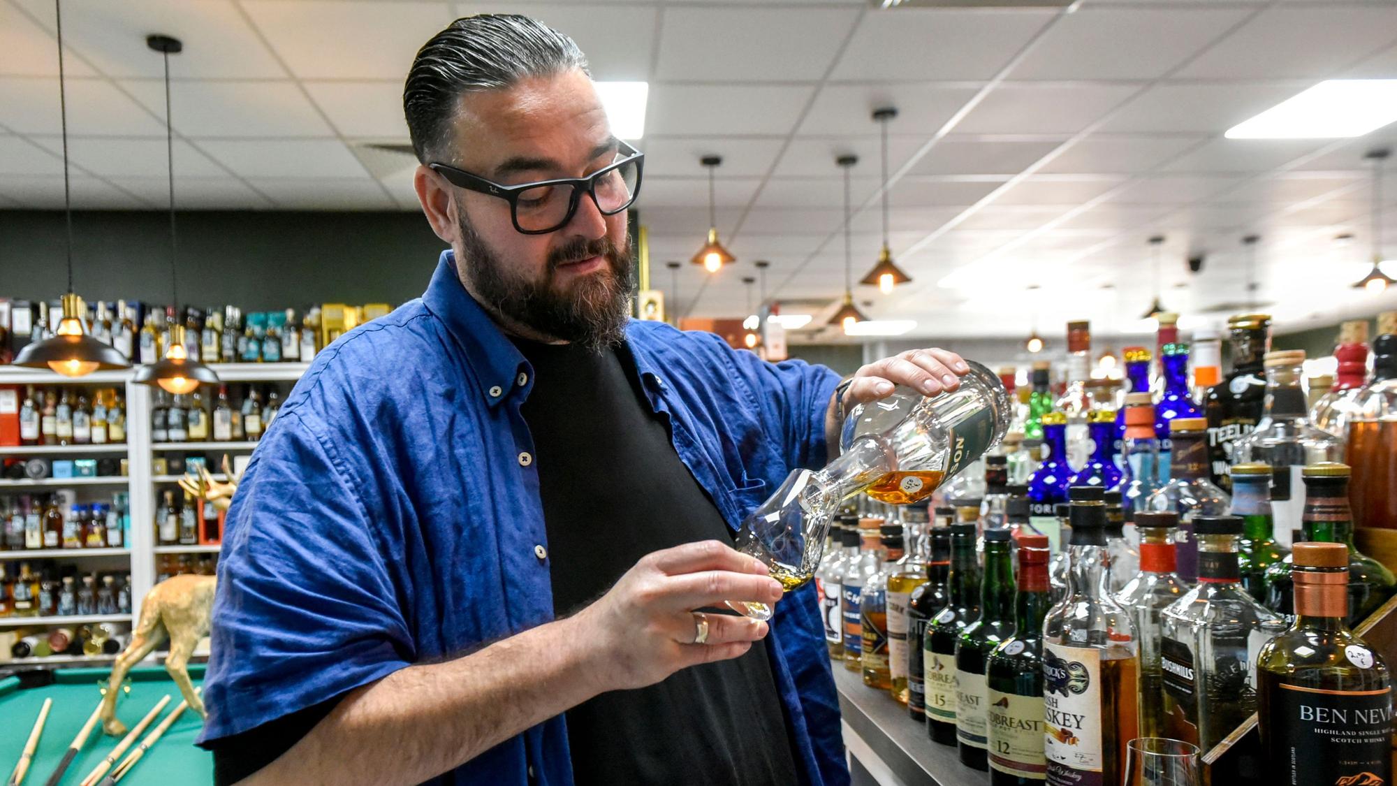 Marco Bonn trägt ein schwarzes T-Shirt und ein blaues Overshirt. Neben ihm ein Regal voller Whiskyflaschen. Das Bild zeigt, wie er ein Glas mit Whisky füllt.
