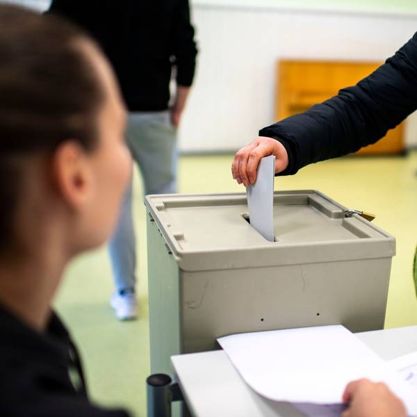 Das Symbolbild zeigt eine Hand, die einen Umschlag in eine Wahlurne wirft. Links im Vordergrund ist unscharf ein Mensch zu sehen.