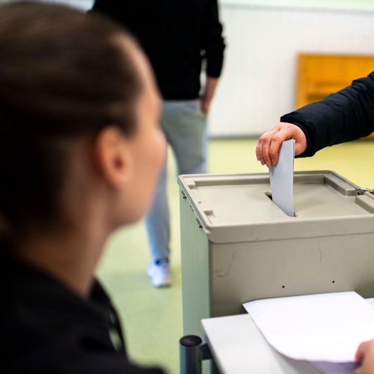 Das Symbolbild zeigt eine Hand, die einen Umschlag in eine Wahlurne wirft. Links im Vordergrund ist unscharf ein Mensch zu sehen.