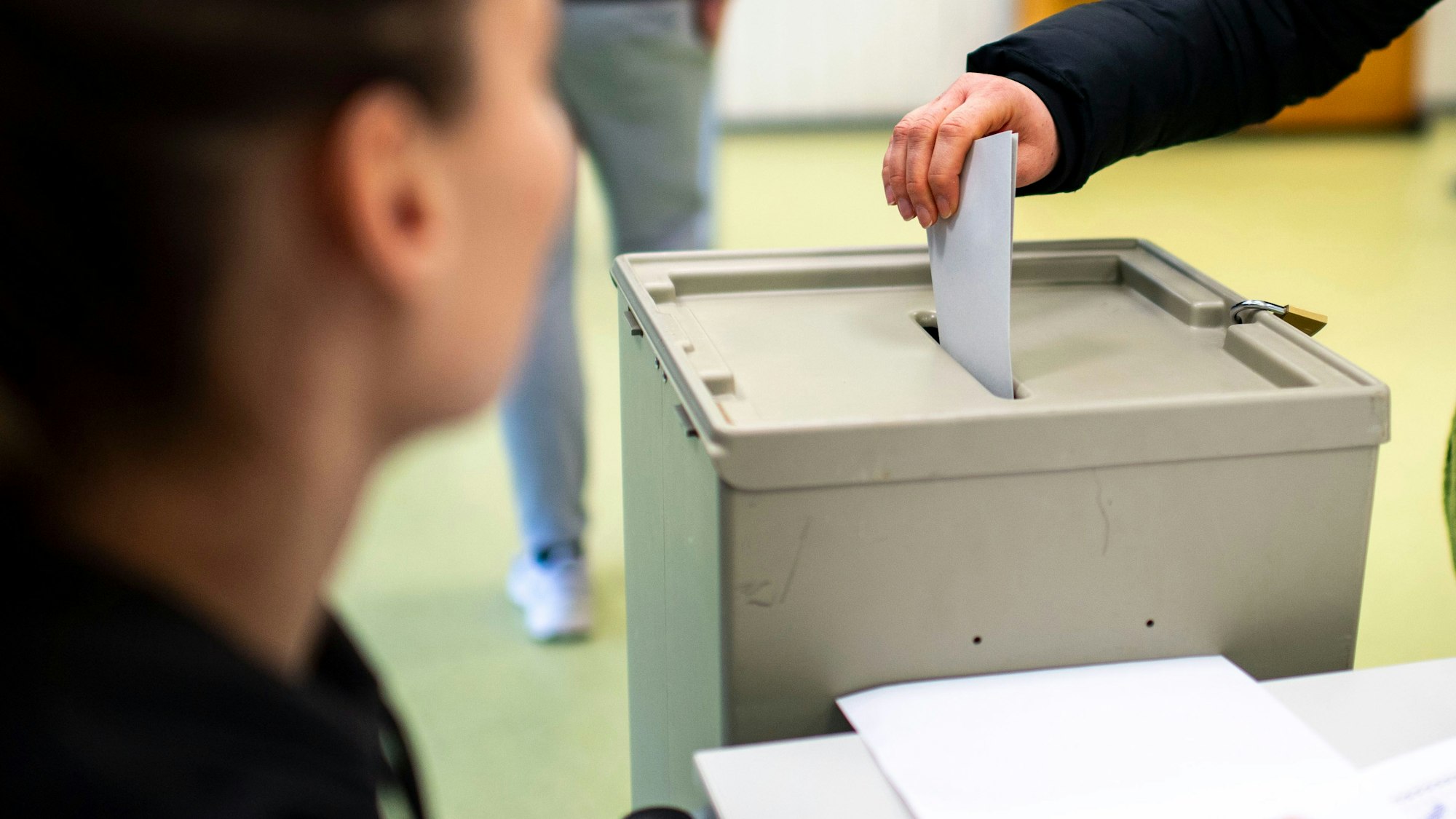 Das Symbolbild zeigt eine Hand, die einen Umschlag in eine Wahlurne wirft. Links im Vordergrund ist unscharf ein Mensch zu sehen.