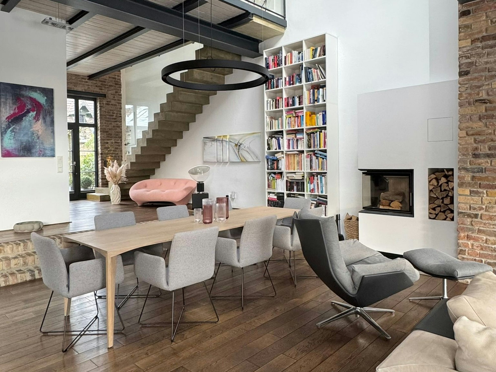 Aufnahme eines Innenbereichs mit gehobener Ausstattung: ein Kamin, aufeinander angepasste Möbel, eine lange Tafel, ein hohes Bücherregal. Der Raum öffnet sich nach oben zu einer Galerie.