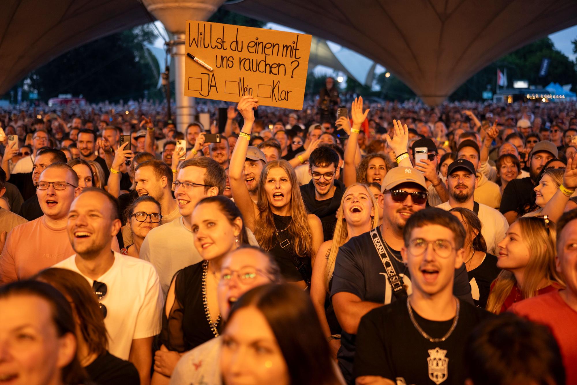 Ein Blick ins Publikum. Eine Frau hält ein Schild hoch: "Willst du einen mit uns rauchen?"