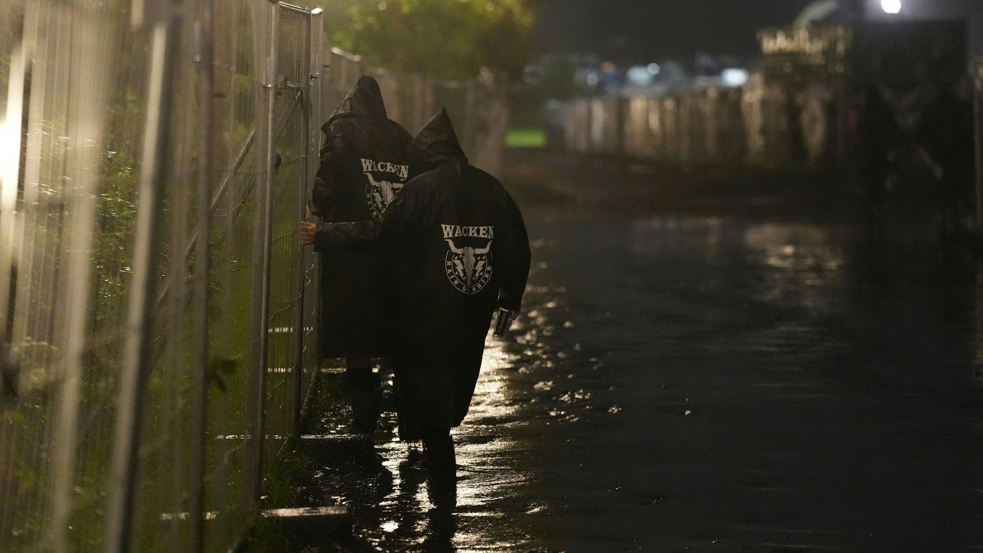 Metal-Fans gehen in der Nacht über einen überfluteten Weg auf dem Festivalgelände des Wacken Open Air (WOA).
