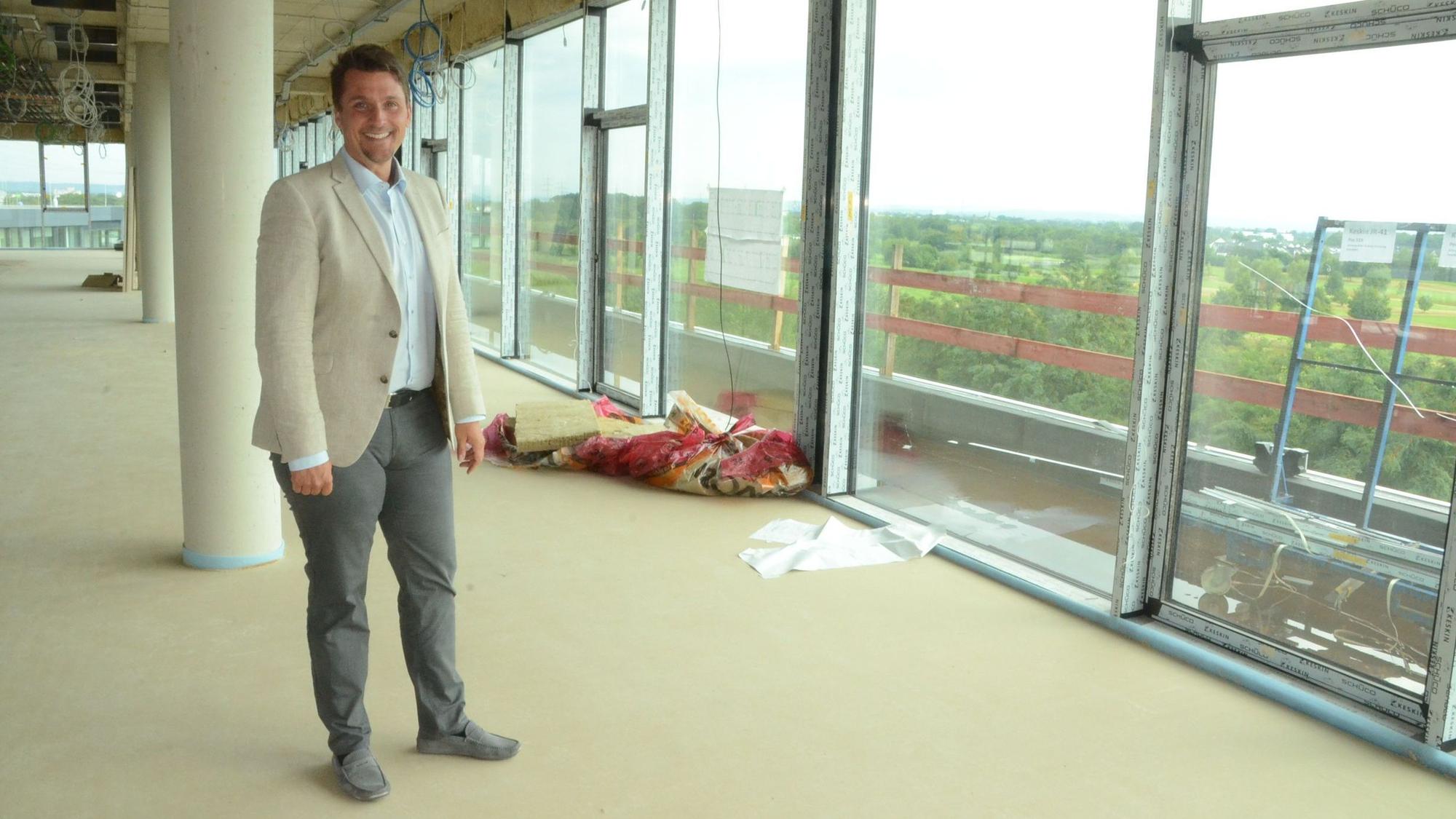 Ein Mann in Hose, Hemd und Sakko in einem noch nicht fertiggestellten Neubau mit großen Fenstern.
