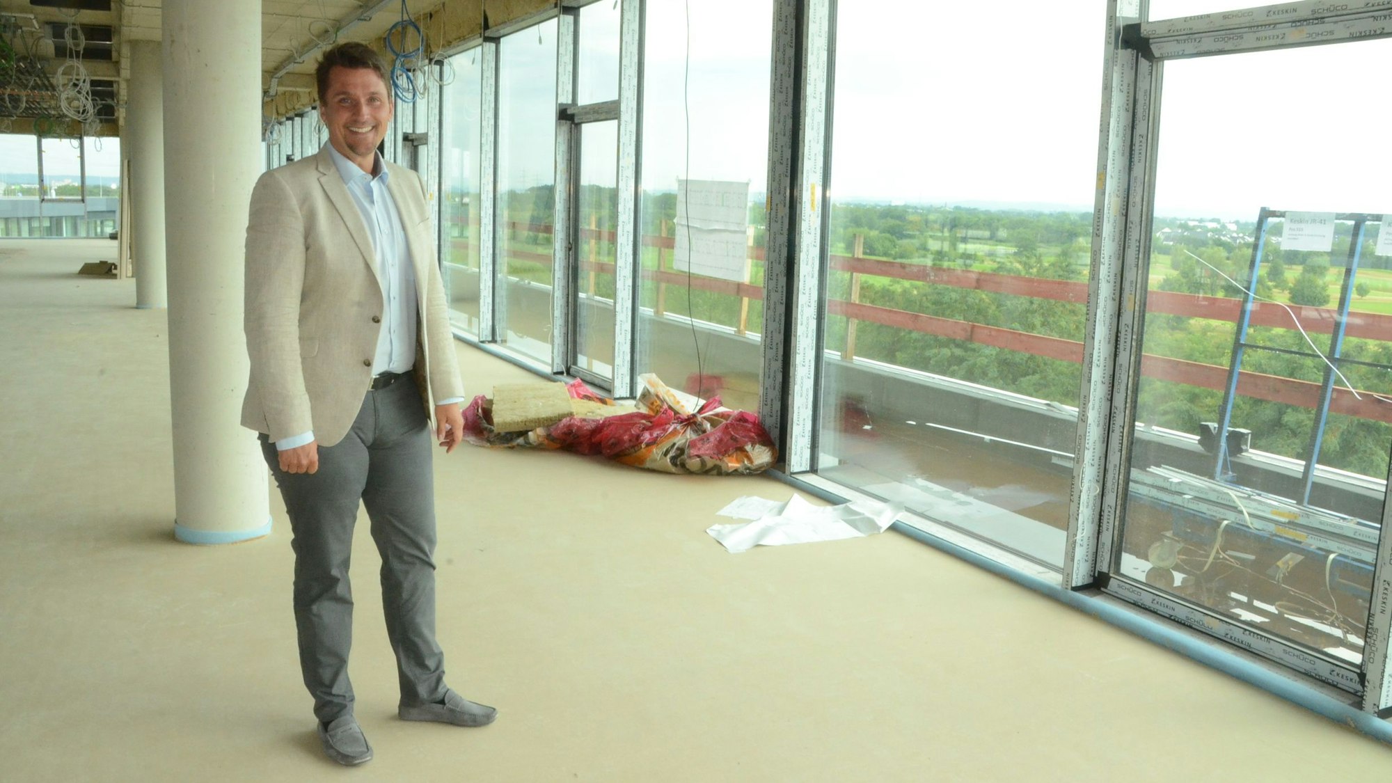Ein Mann in Hose, Hemd und Sakko in einem noch nicht fertiggestellten Neubau mit großen Fenstern.