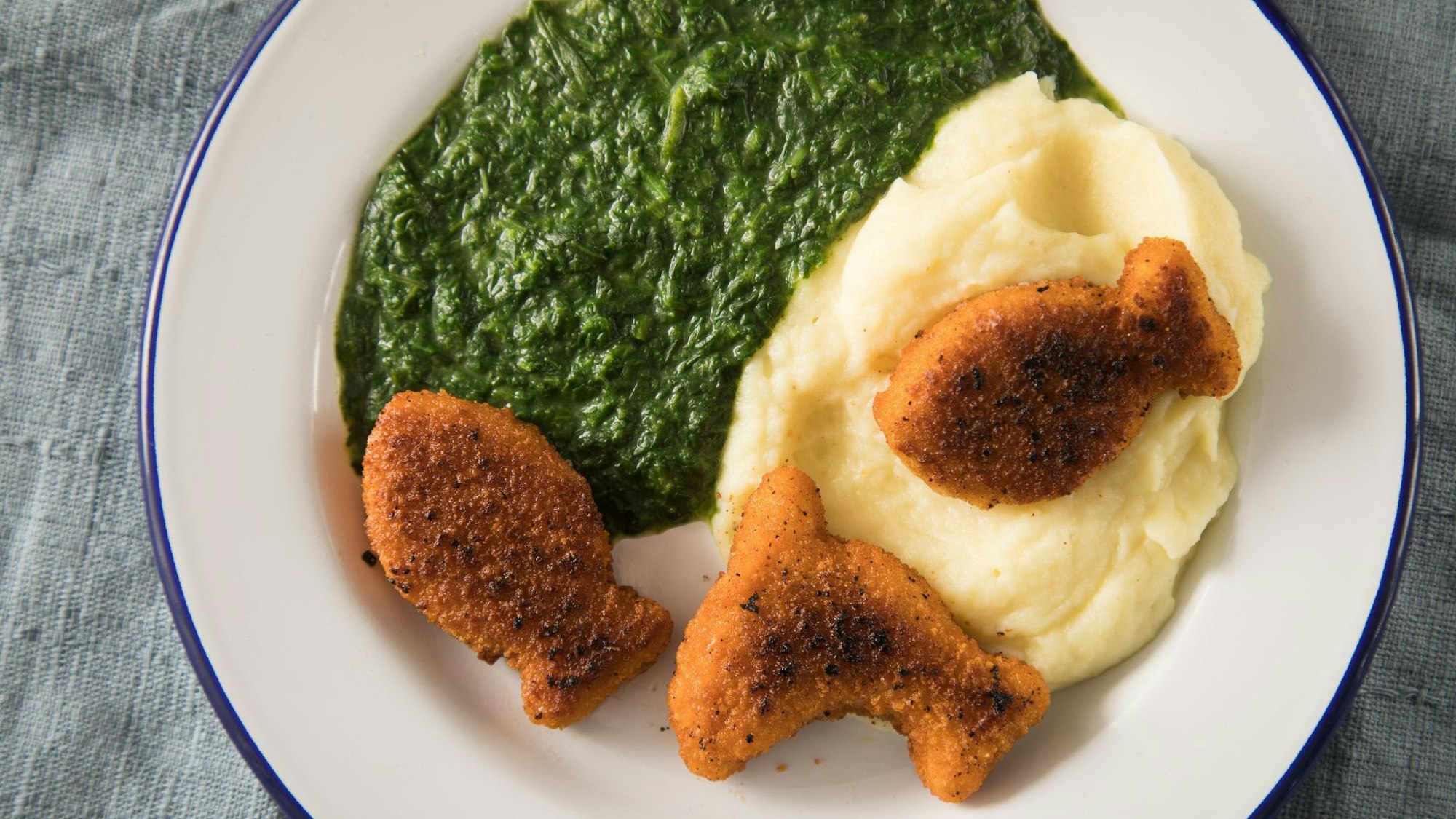Ein Mahl auf einem Teller bestehend aus Spinat, Kartoffelbrei und Fischnuggets.