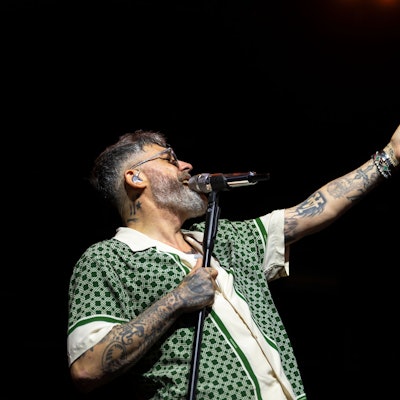 Sido gestikuliert am Mikrofon mit ausgestrecktem Arm und geöffneter Hand. Er trägt ein grün-gemustertes Hemd.