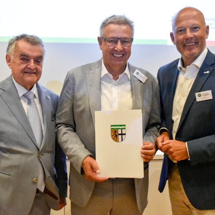 Das Bild zeigt drei Männer, einer hält eine Urkunde in seinen Händen.