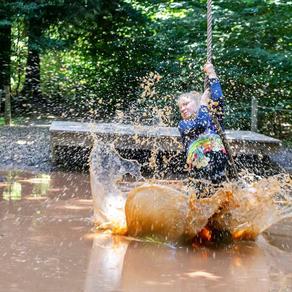 Ein Mädchen schwingt sich mit einem Tau über ein Becken mit matschigem Wasser. Mit den Füßen wirbelt es das Wasser auf, das hochspritzt.
