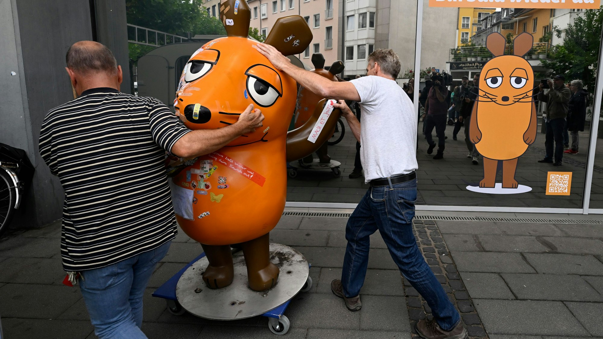 Zwei Personen schieben eine große Maus-Figur auf einem Rollbrett.