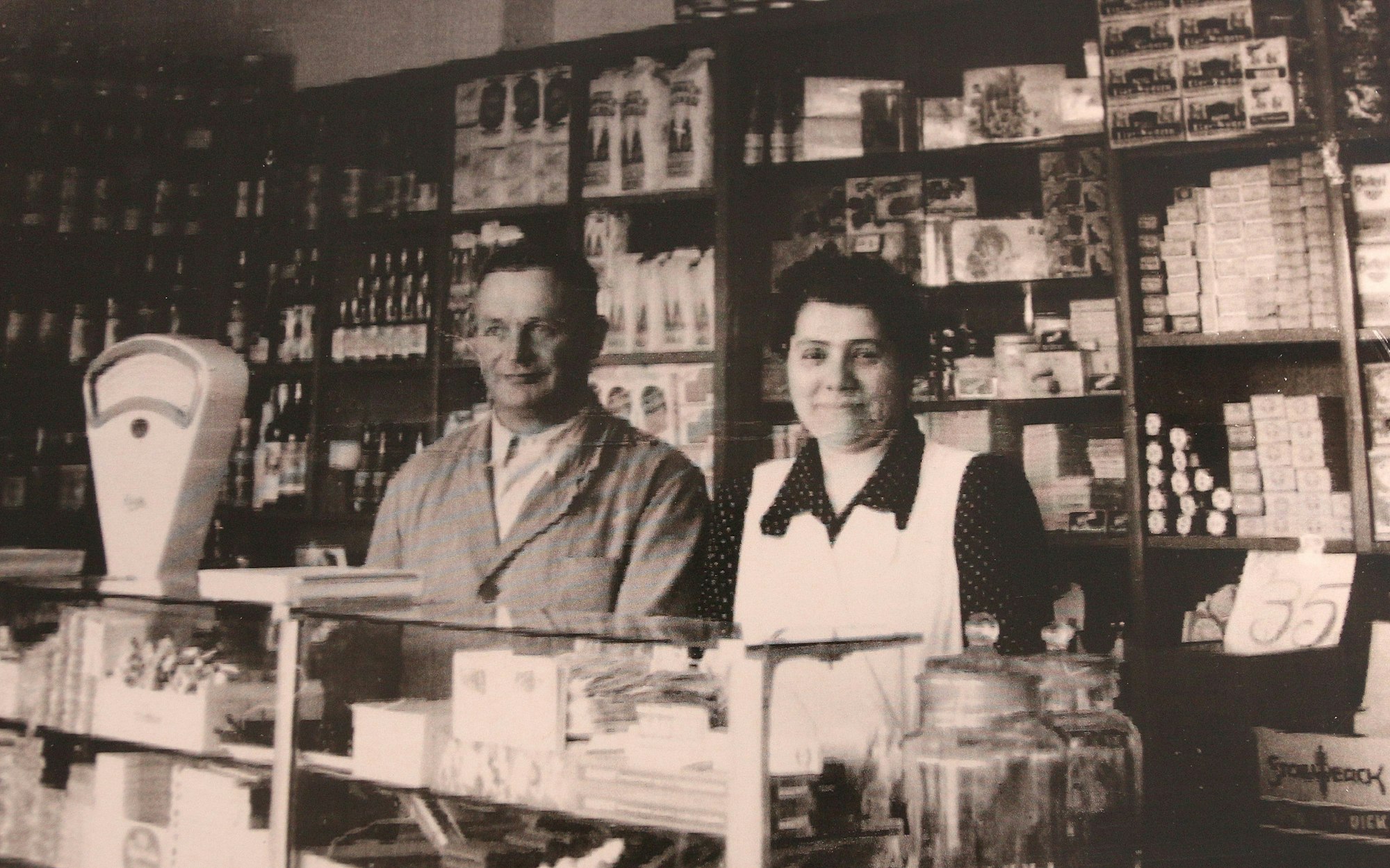 Das Bild zeigt den Laden im Jahr 1930. Das Bild ist schwarz-weiß. Das Ehepaar steht an der Kasse, hinter ihnen viele Waren in Schränken.