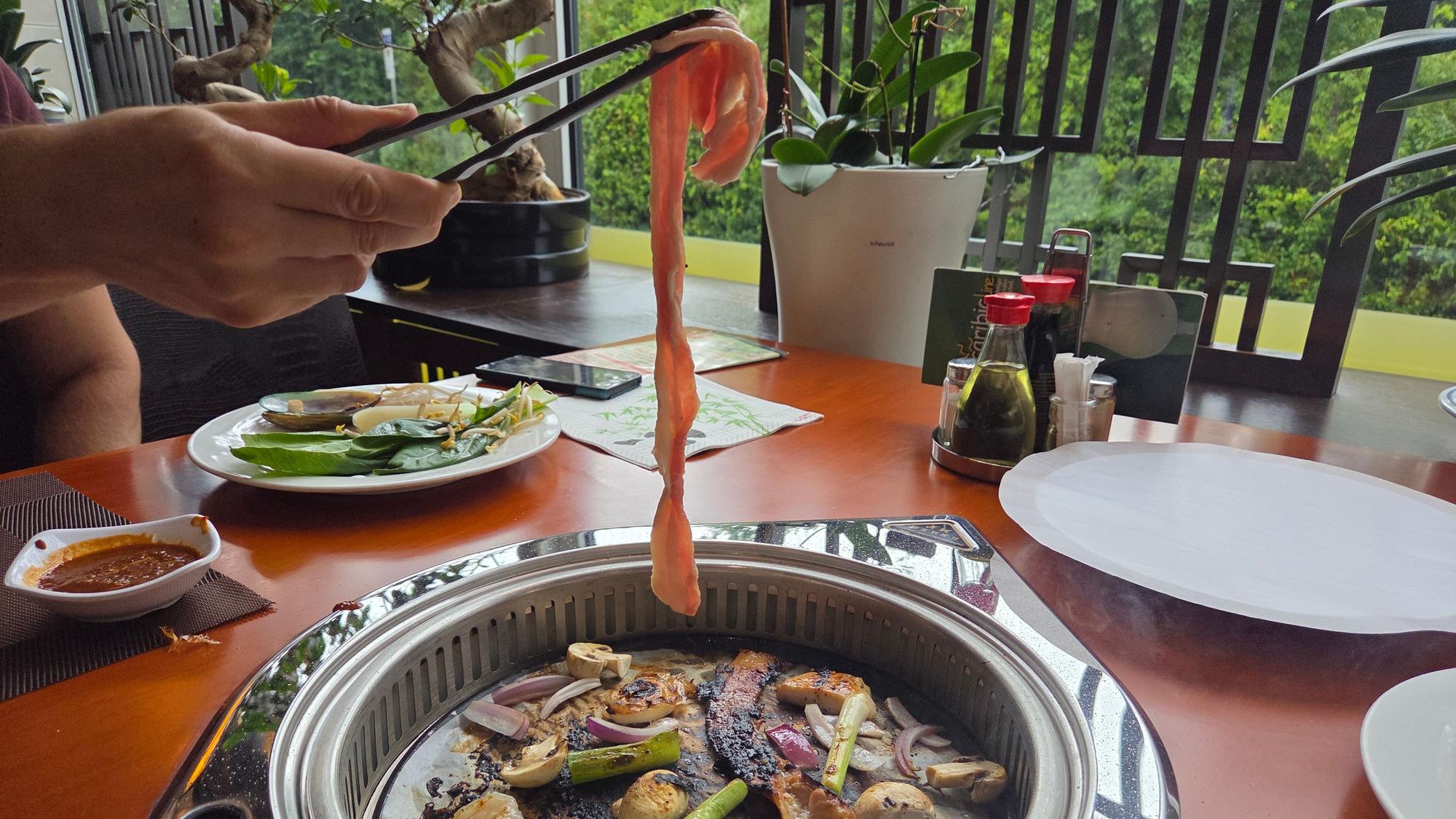 Eine Person hebt mit einer Zange eine Scheibe Schweinebauch auf den Grill.