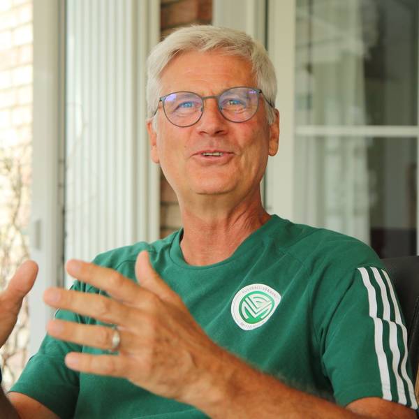 Uwe Stark sitzt gestikulierend und lachend auf einem Stuhl. Er trägt ein offizielles T-Shirt mit FVM-Logo.