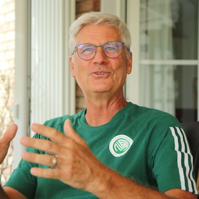 Uwe Stark sitzt gestikulierend und lachend auf einem Stuhl. Er trägt ein offizielles T-Shirt mit FVM-Logo.