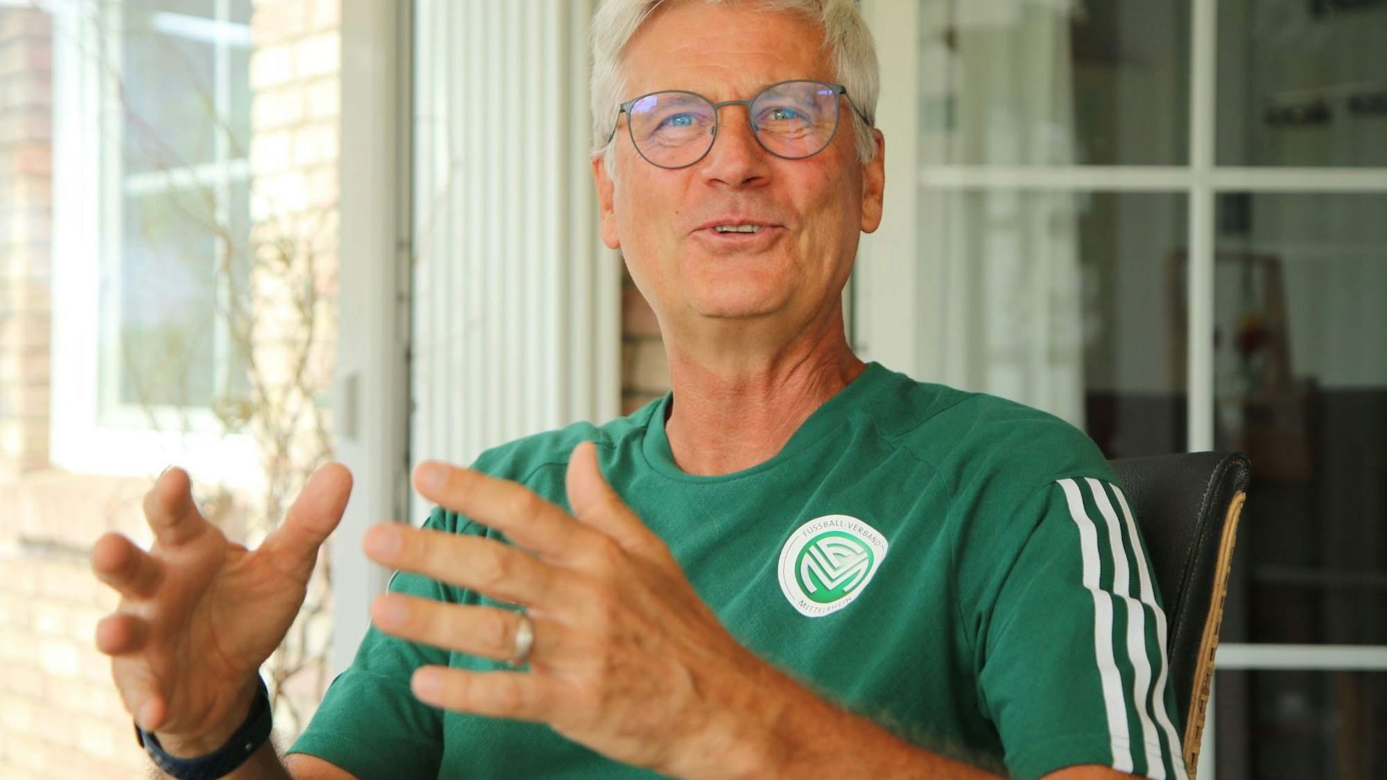 Uwe Stark sitzt gestikulierend und lachend auf einem Stuhl. Er trägt ein offizielles T-Shirt mit FVM-Logo.