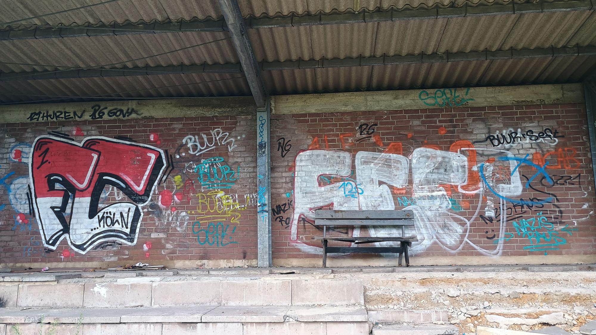 Das Bild zeigt die Wand hinter der Tribüne. Sie ist voller Graffiti. Eine einzelne Bank steht an der Wand.
