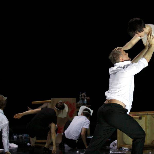 Ein Schauspieler fängt eine Schauspielerin auf, die mit ausgestreckten Beinen von einer Tischkante zu kippen droht.