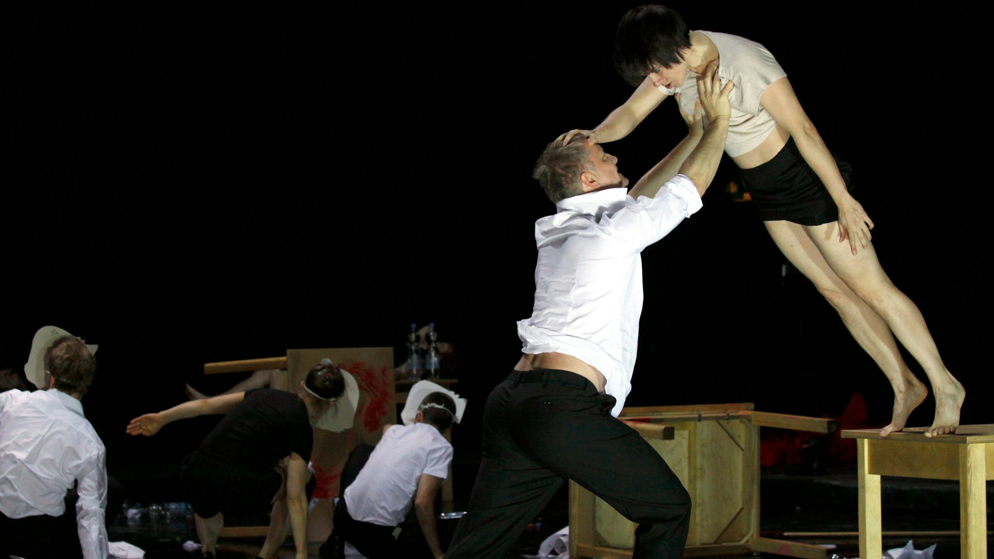 Ein Schauspieler fängt eine Schauspielerin auf, die mit ausgestreckten Beinen von einer Tischkante zu kippen droht.