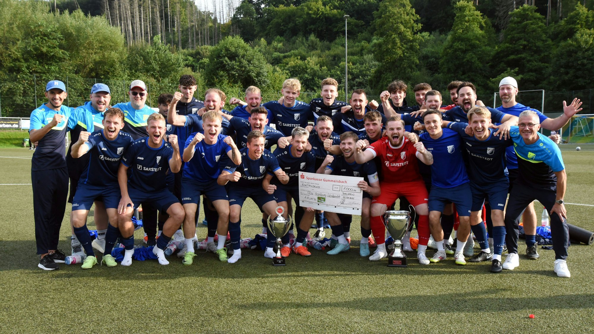 Eine jubelnde Fußballmannschaft mit Pokal und einem Scheck der Sparkasse Gummersbach über 100 Euro.