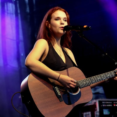 Eine junge Frau steht auf einer Bühne, sie singt und spielt Gitarre.