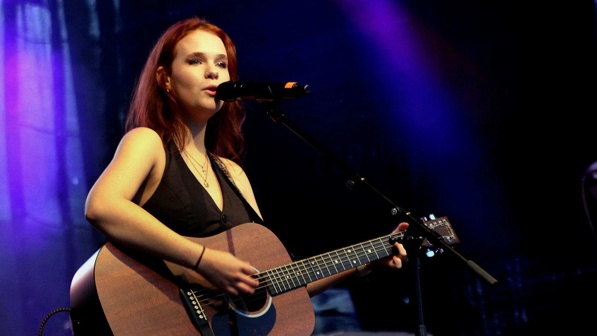 Eine junge Frau steht auf einer Bühne, sie singt und spielt Gitarre.
