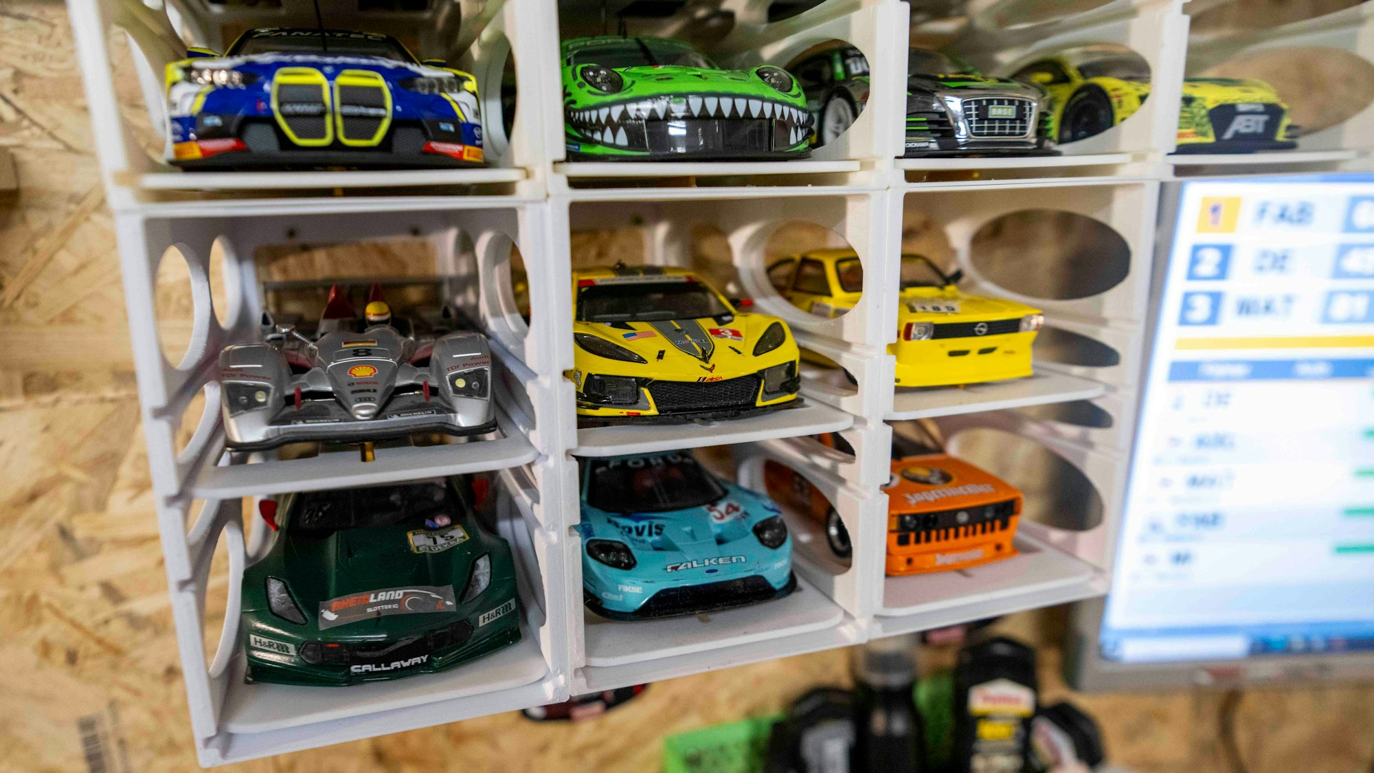 Zehn verschiedene Carrera-Rennwagen im Maßstab 1:24 parken in ihren Plastikgaragen vor einer OSB-Wand.