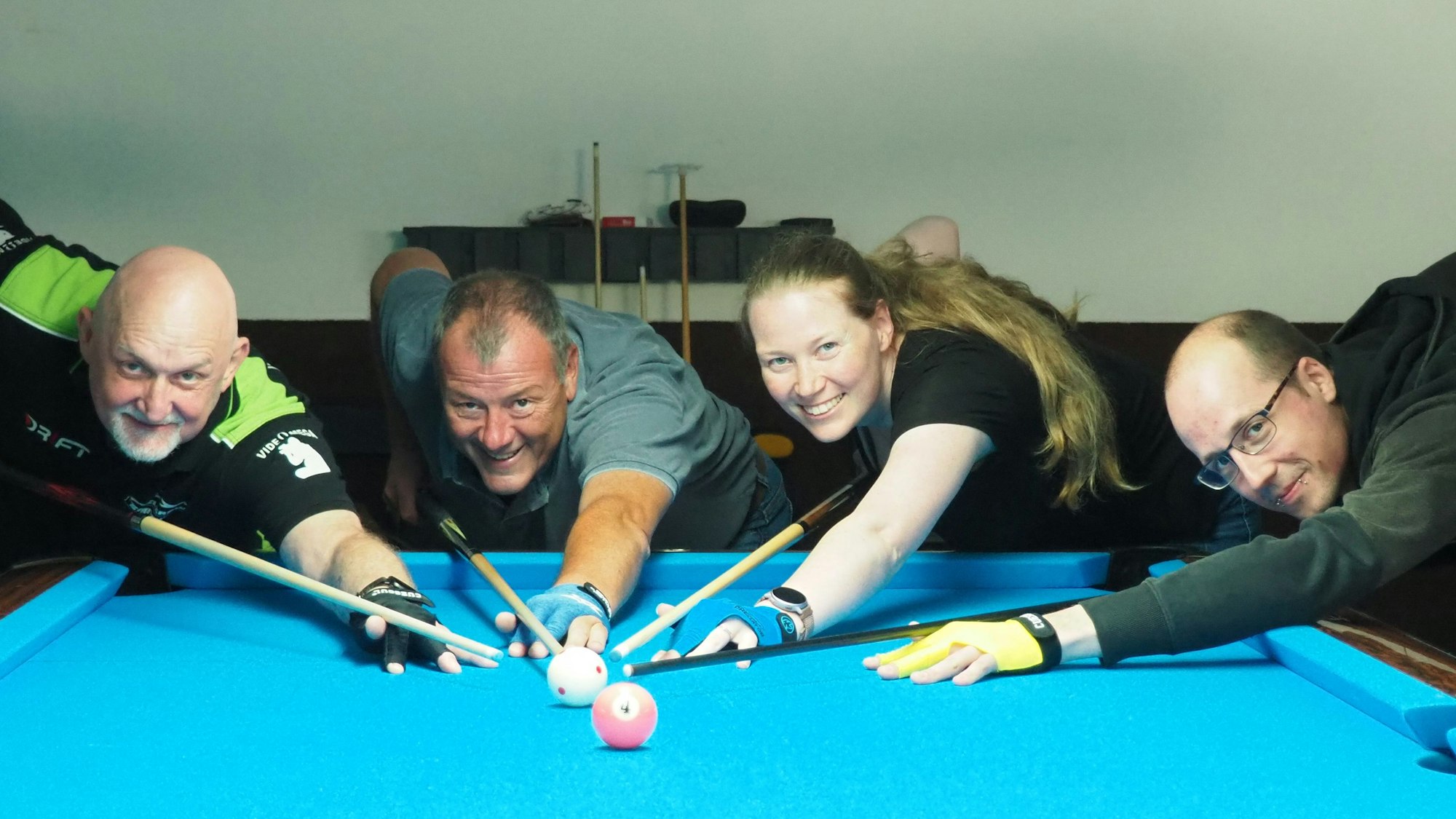 Ritchy Ohm (v.l.), Larissa Henig, Lothar Lubitz und Nick Henig zielen an einem blau bespannten Billardtisch mit ihren Queues auf eine weiße Kugel.