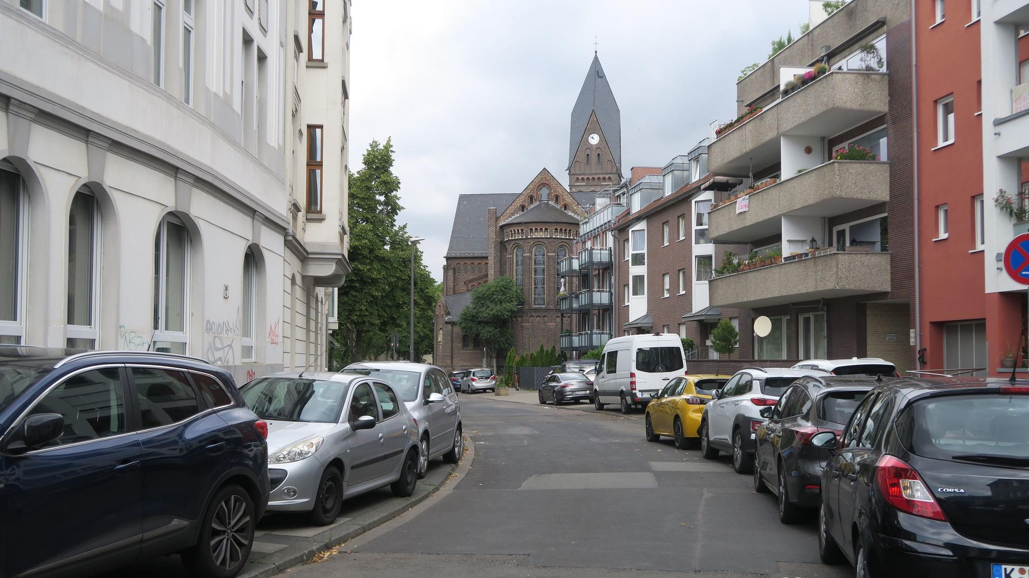 Am Straßenrand einer etwas geflickten Straße stehen viele Autos, teils auf der Straße, teils auf dem Bürgersteig.