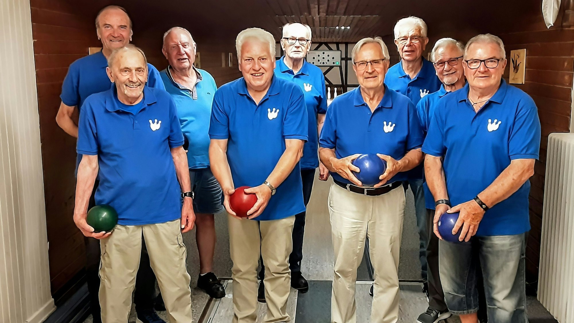 Acht ältere Herren mit blauen Vereinshemden, auf denen Kegel zu sehen sind, stehen auf einer Kegelbahn.