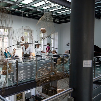 Menschen, die die Ausstellung besuchen, stehen und sitzen vor Kunstinstallationen, unter anderem zusammengedrückten Vogelkäfigen.