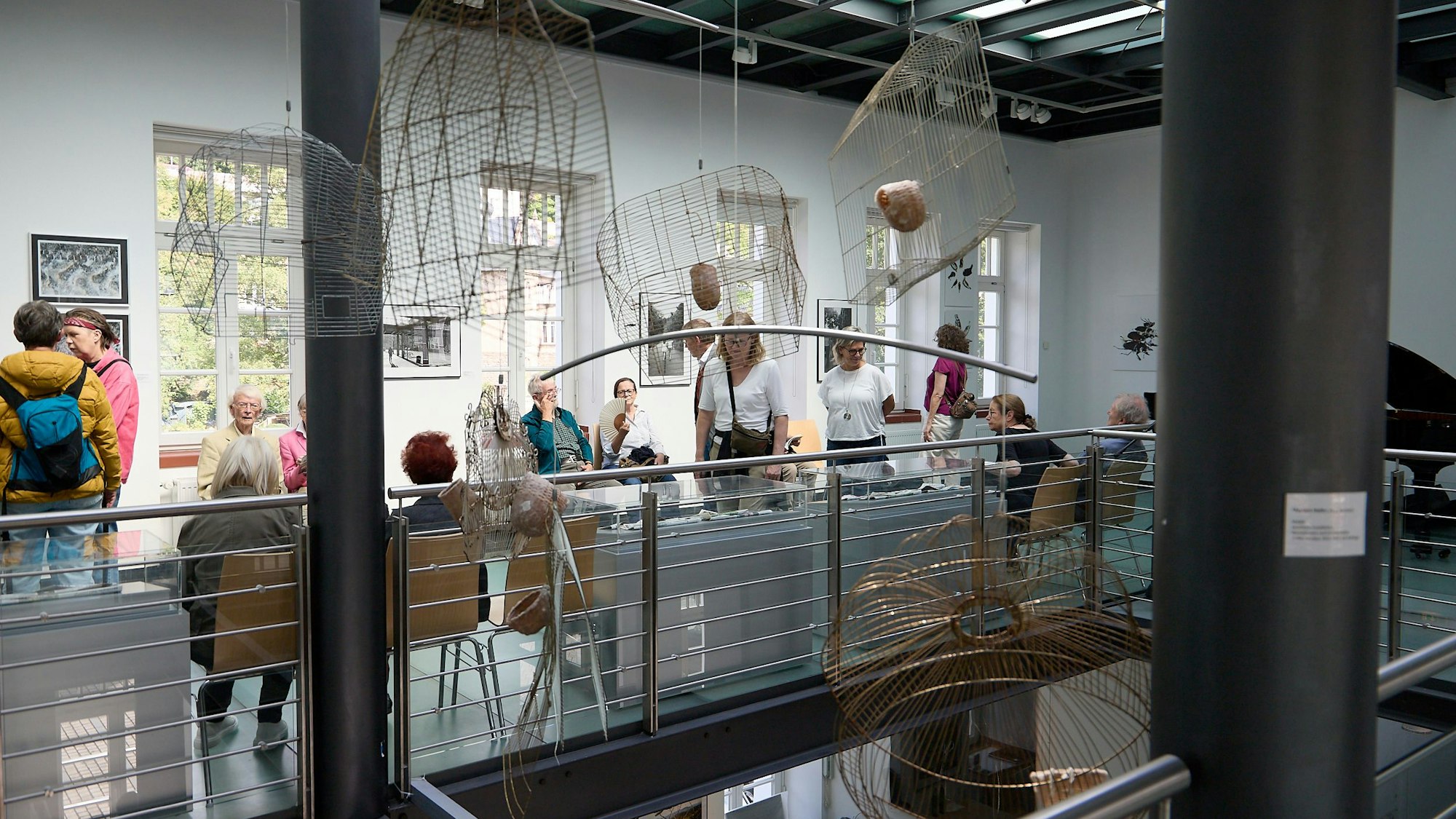 Menschen, die die Ausstellung besuchen, stehen und sitzen vor Kunstinstallationen, unter anderem zusammengedrückten Vogelkäfigen.