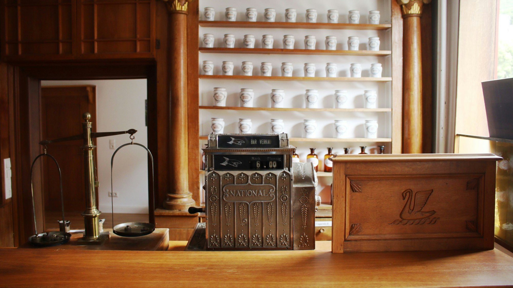 Aus dem Verkaufsraum gut sichtbar sind von links eine Waage, eine Kasse und eine Holztafel, auf der ein Schwan abgebildet ist.