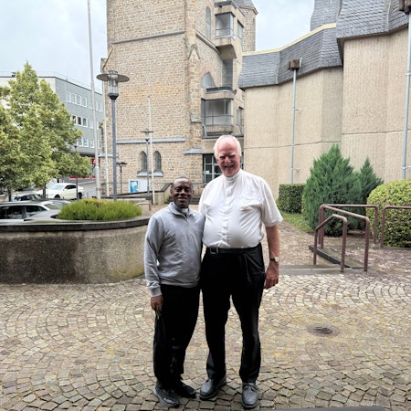 Pater Domisias Kayemba und Kreisdechant Christoph Bersch stehen vor einer Kirche nebeneinander