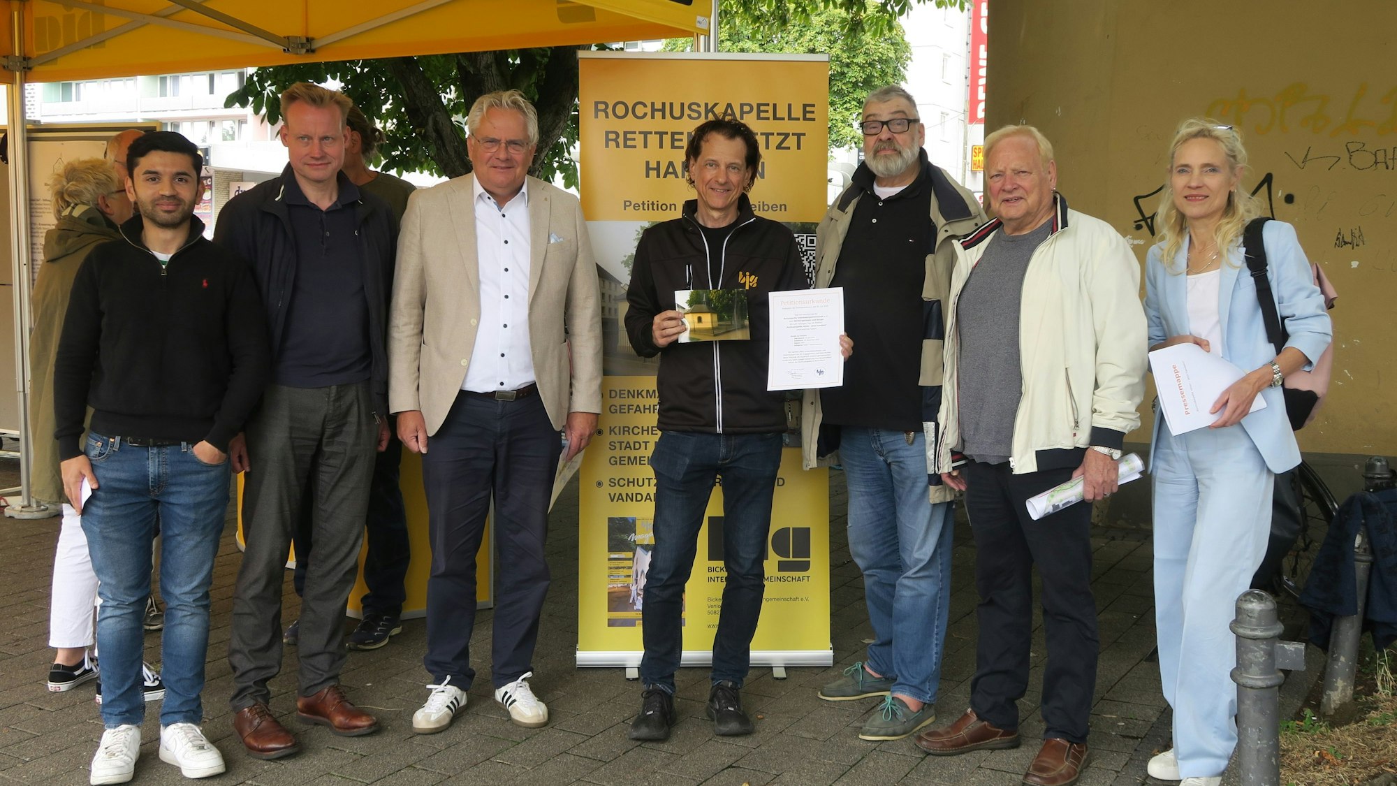 Sieben Menschen stehen vor einem Aufsteller unter einem Zeltdach.